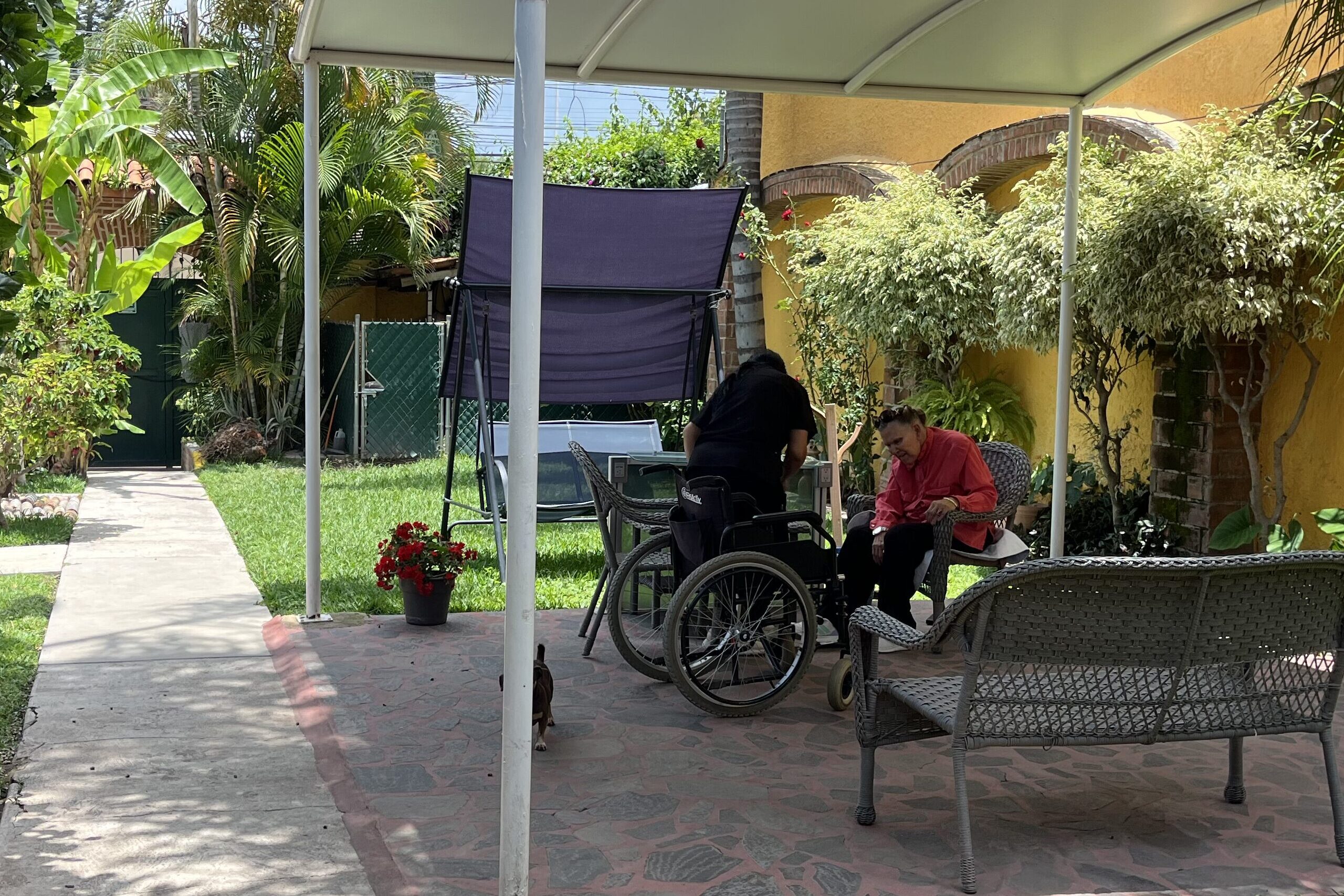 Caregiver assisting a resident in a wheelchair on the garden patio at Rudy & Magda's care home in Mexico
